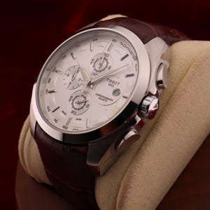 Puede incluir: Un reloj de cuarzo cron&oacute;grafo Tissot 1853 plateado con esfera blanca y correa de cuero marr&oacute;n. El reloj presenta m&uacute;ltiples subesferas y una escala taquim&eacute;trica. El reloj se exhibe sobre un coj&iacute;n beige sobre un fondo rojo oscuro.
