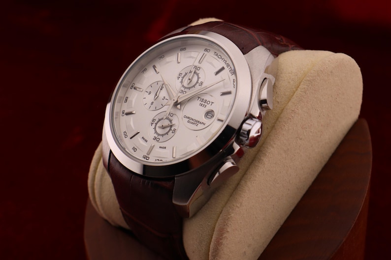 Puede incluir: Un reloj de cuarzo cron&oacute;grafo Tissot 1853 plateado con esfera blanca y correa de cuero marr&oacute;n. El reloj presenta m&uacute;ltiples subesferas y una escala taquim&eacute;trica. El reloj se exhibe sobre un coj&iacute;n beige sobre un fondo rojo oscuro.
