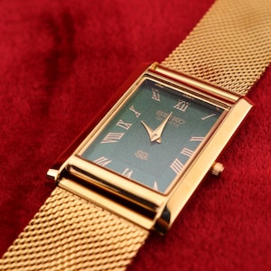 K&ouml;nnte beinhalten: Rechteckige Seiko Quartz-Uhr in Goldoptik mit einem Mesh-Armband. Das Zifferblatt der Uhr ist tiefgr&uuml;n mit goldenen r&ouml;mischen Ziffern und Zeigern. Die Uhr befindet sich vor einem roten Hintergrund.