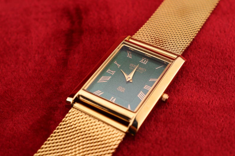K&ouml;nnte beinhalten: Rechteckige Seiko Quartz-Uhr in Goldoptik mit einem Mesh-Armband. Das Zifferblatt der Uhr ist tiefgr&uuml;n mit goldenen r&ouml;mischen Ziffern und Zeigern. Die Uhr befindet sich vor einem roten Hintergrund.