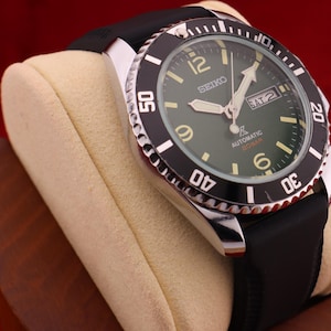 Può includere: Un orologio subacqueo automatico Seiko con lunetta nera e quadrante verde. L'orologio ha un cinturino in gomma nero ed è esposto su un cuscino per orologi color crema. Il quadrante mostra il giorno e la data.