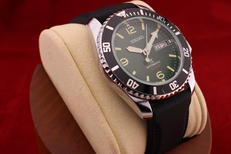 Puede incluir: Un reloj de buceo autom&aacute;tico Seiko con bisel negro y esfera verde. El reloj tiene una correa de caucho negra y se exhibe sobre una almohada para reloj color crema. La esfera muestra el d&iacute;a y la fecha.