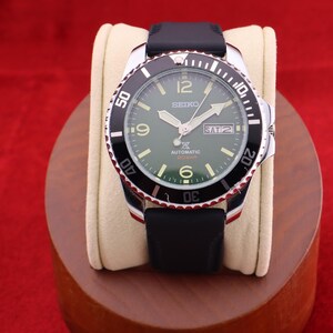 Puede incluir: Un reloj autom&aacute;tico Seiko con bisel negro y plateado, correa negra y esfera verde. El reloj muestra el d&iacute;a y la fecha, con n&uacute;meros y agujas blancas. El reloj se exhibe sobre un coj&iacute;n color crema.