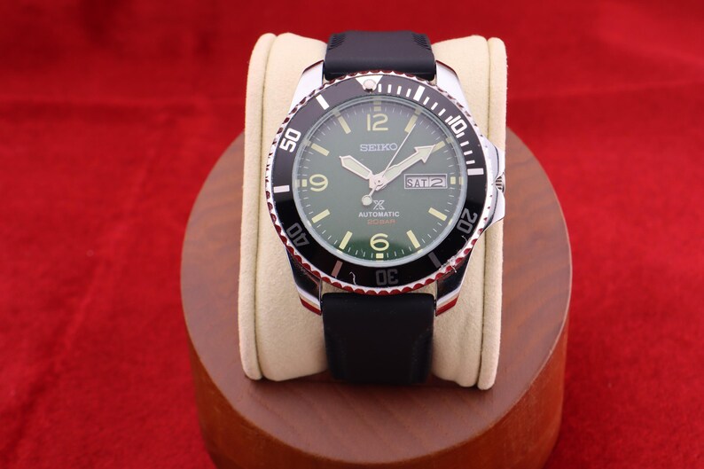 Puede incluir: Un reloj autom&aacute;tico Seiko con bisel negro y plateado, correa negra y esfera verde. El reloj muestra el d&iacute;a y la fecha, con n&uacute;meros y agujas blancas. El reloj se exhibe sobre un coj&iacute;n color crema.
