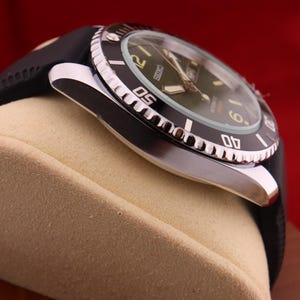 Puede incluir: Primer plano de un reloj de pulsera negro y plateado con una correa de goma negra. La esfera del reloj es verde con n&uacute;meros y agujas blancas. El bisel es negro con marcas blancas. El reloj se muestra sobre una superficie beige sobre un fondo rojo.