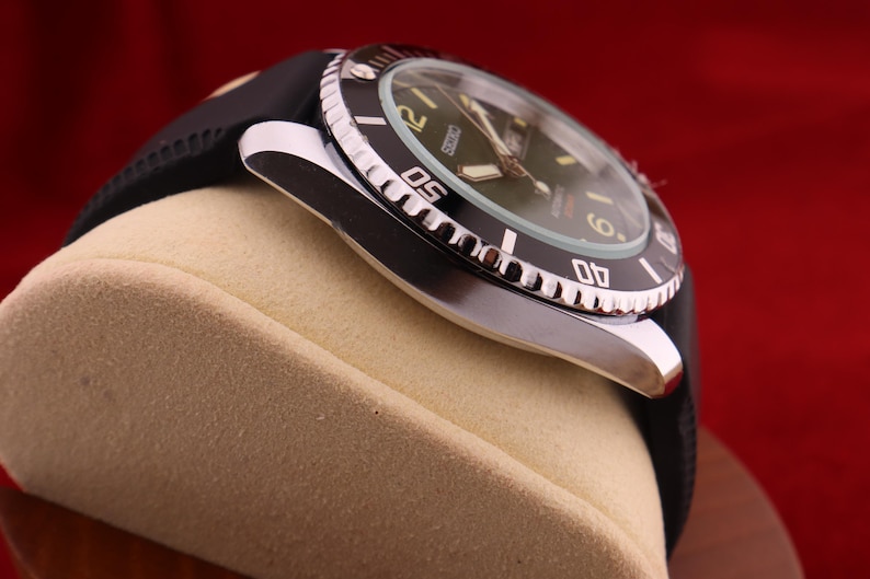 Puede incluir: Primer plano de un reloj de pulsera negro y plateado con una correa de goma negra. La esfera del reloj es verde con n&uacute;meros y agujas blancas. El bisel es negro con marcas blancas. El reloj se muestra sobre una superficie beige sobre un fondo rojo.