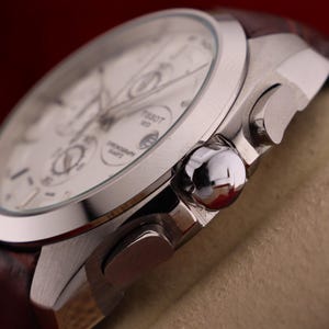 Puede incluir: Primer plano de un reloj cron&oacute;grafo Tissot plateado con esfera blanca y correa de cuero marr&oacute;n. El reloj presenta m&uacute;ltiples subesferas y pulsadores. El reloj se muestra sobre un fondo neutro.