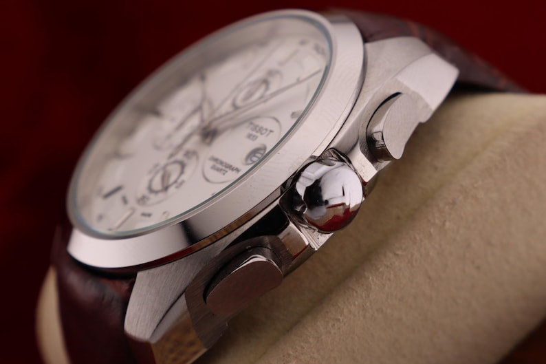 Puede incluir: Primer plano de un reloj cron&oacute;grafo Tissot plateado con esfera blanca y correa de cuero marr&oacute;n. El reloj presenta m&uacute;ltiples subesferas y pulsadores. El reloj se muestra sobre un fondo neutro.