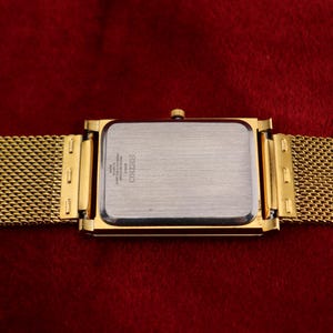K&ouml;nnte beinhalten: Goldfarbene rechteckige Uhr mit einem Mesh-Armband. Das Zifferblatt der Uhr ist silberfarben mit schwarzem Text. Das Armband und das Geh&auml;use sind goldfarben. Die Uhr befindet sich vor einem tiefroten Hintergrund.