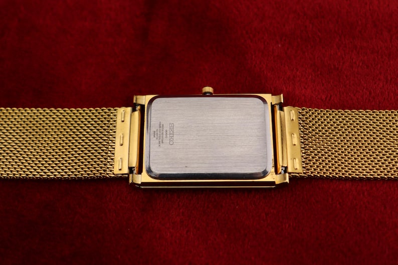 K&ouml;nnte beinhalten: Goldfarbene rechteckige Uhr mit einem Mesh-Armband. Das Zifferblatt der Uhr ist silberfarben mit schwarzem Text. Das Armband und das Geh&auml;use sind goldfarben. Die Uhr befindet sich vor einem tiefroten Hintergrund.