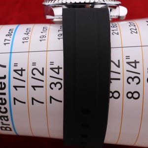 Puede incluir: Una correa de reloj negra est&aacute; envuelta alrededor de una tabla de medici&oacute;n de pulseras. La tabla muestra medidas en pulgadas y cent&iacute;metros, que van desde 6 1/2 pulgadas (16,5 cm) hasta 9 pulgadas (22,8 cm). El reloj tiene un bisel plateado.