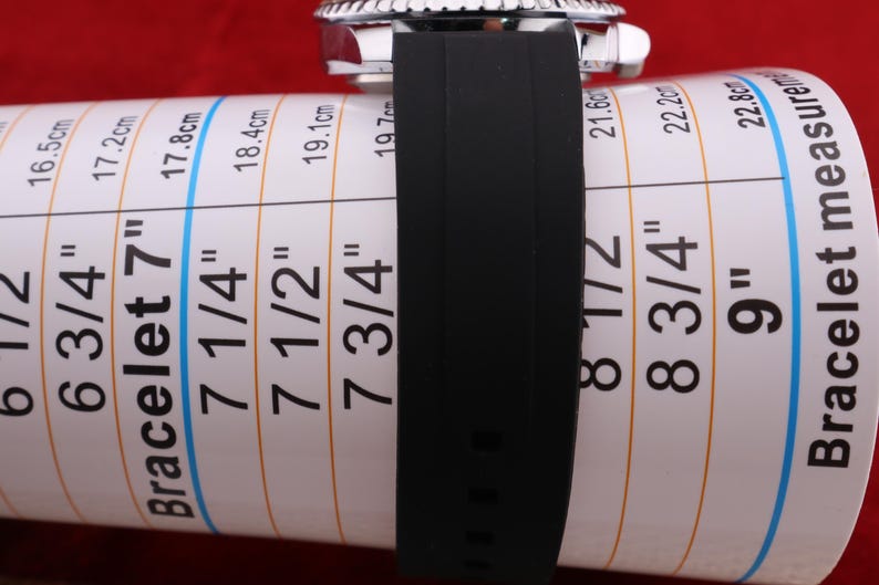 Puede incluir: Una correa de reloj negra est&aacute; envuelta alrededor de una tabla de medici&oacute;n de pulseras. La tabla muestra medidas en pulgadas y cent&iacute;metros, que van desde 6 1/2 pulgadas (16,5 cm) hasta 9 pulgadas (22,8 cm). El reloj tiene un bisel plateado.
