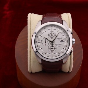 Puede incluir: Reloj cron&oacute;grafo Tissot 1853 plateado con correa de cuero marr&oacute;n. El reloj tiene una esfera blanca con m&uacute;ltiples subesferas y una ventana de fecha. El reloj se exhibe sobre un coj&iacute;n crema y un soporte de madera.