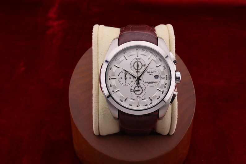 Puede incluir: Reloj cron&oacute;grafo Tissot 1853 plateado con correa de cuero marr&oacute;n. El reloj tiene una esfera blanca con m&uacute;ltiples subesferas y una ventana de fecha. El reloj se exhibe sobre un coj&iacute;n crema y un soporte de madera.
