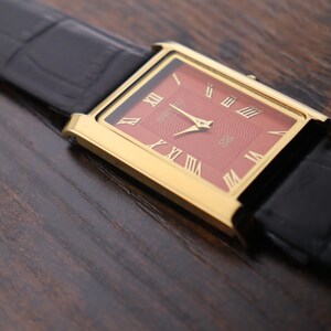Op de afbeelding: Een gouden rechthoekig polshorloge met een rode wijzerplaat en gouden Romeinse cijfers. Het horloge heeft gouden wijzers en een zwarte leren band. Het horloge wordt getoond op een donker houten oppervlak.