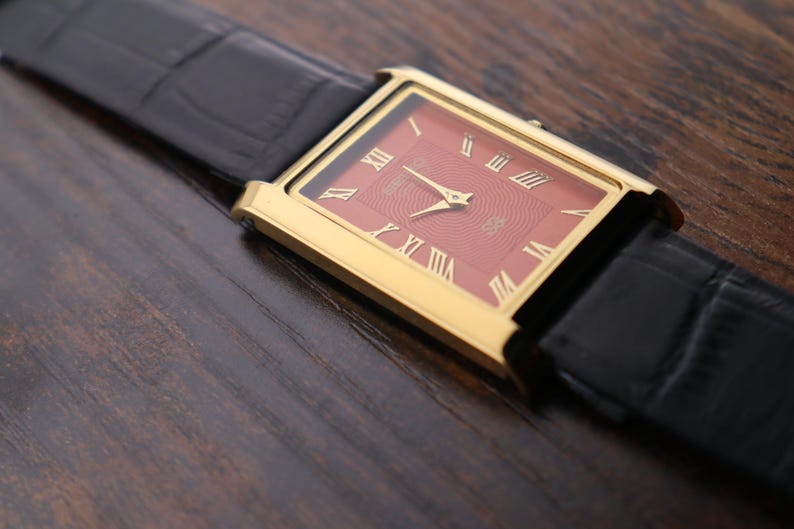 Op de afbeelding: Een gouden rechthoekig polshorloge met een rode wijzerplaat en gouden Romeinse cijfers. Het horloge heeft gouden wijzers en een zwarte leren band. Het horloge wordt getoond op een donker houten oppervlak.
