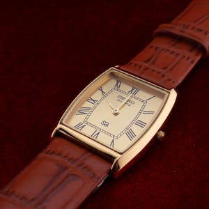 May include: A gold-toned rectangular Seiko Quartz watch with a light-colored face and Roman numeral hour markers. The watch has a brown leather band. The watch is displayed on a dark red surface.