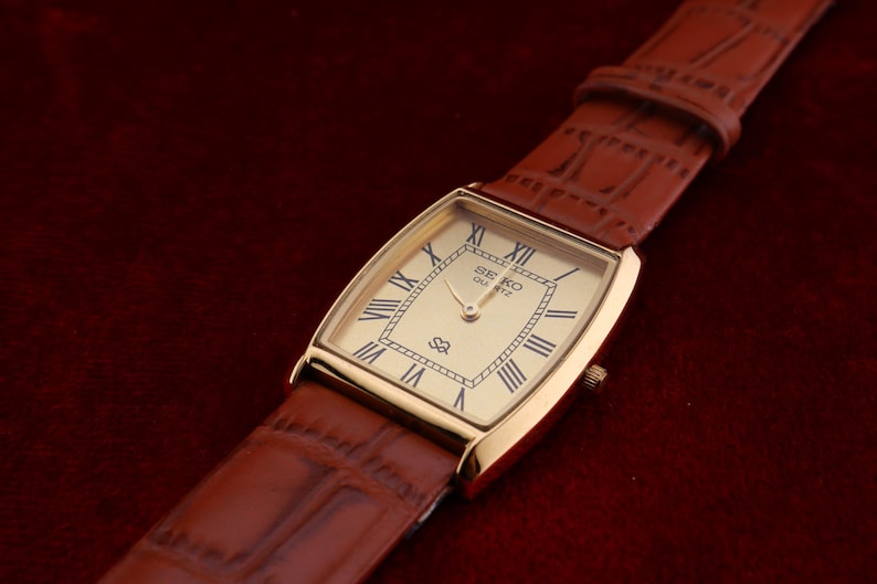 May include: A gold-toned rectangular Seiko Quartz watch with a light-colored face and Roman numeral hour markers. The watch has a brown leather band. The watch is displayed on a dark red surface.