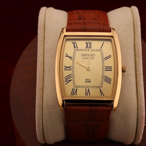 May include: A gold-toned Seiko Quartz watch with a rectangular face and Roman numeral hour markers. The watch has a light-colored dial and a brown leather band. The watch is displayed on a cream-colored cushion.