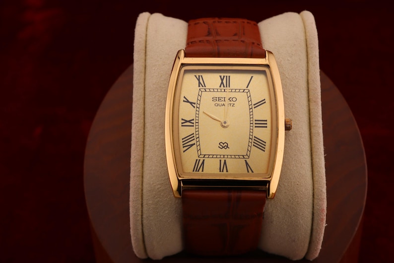May include: A gold-toned Seiko Quartz watch with a rectangular face and Roman numeral hour markers. The watch has a light-colored dial and a brown leather band. The watch is displayed on a cream-colored cushion.