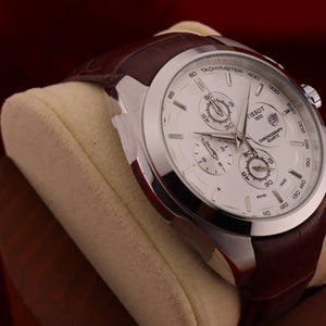 Puede incluir: Un reloj cron&oacute;grafo Tissot plateado con esfera blanca y tres subesferas. El reloj tiene una correa de cuero marr&oacute;n y se exhibe sobre un coj&iacute;n color crema. La esfera del reloj tiene las palabras "Tissot" y "1853".