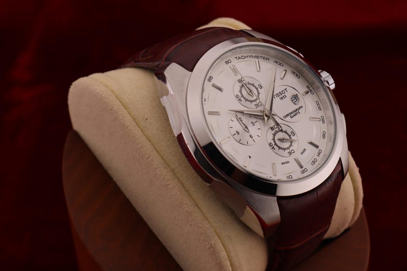 Puede incluir: Un reloj cron&oacute;grafo Tissot plateado con esfera blanca y tres subesferas. El reloj tiene una correa de cuero marr&oacute;n y se exhibe sobre un coj&iacute;n color crema. La esfera del reloj tiene las palabras "Tissot" y "1853".