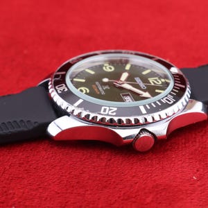 Puede incluir: Un reloj de pulsera plateado con bisel negro y correa de goma negra. La esfera del reloj es verde oscuro con n&uacute;meros y agujas blancos. El reloj est&aacute; sobre un fondo rojo.