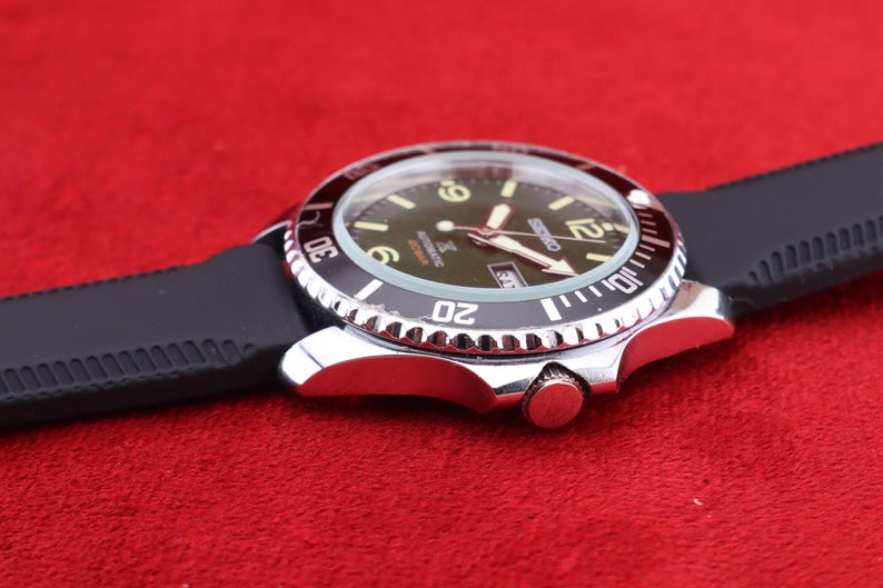 Puede incluir: Un reloj de pulsera plateado con bisel negro y correa de goma negra. La esfera del reloj es verde oscuro con n&uacute;meros y agujas blancos. El reloj est&aacute; sobre un fondo rojo.