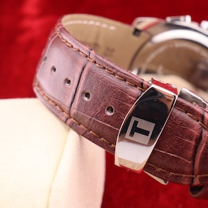 Puede incluir: Primer plano de una correa de reloj de cuero marr&oacute;n con una hebilla de acero inoxidable. La correa del reloj tiene un patr&oacute;n texturizado, similar al de un cocodrilo. La esfera del reloj es visible en el fondo. La correa del reloj est&aacute; sobre una superficie blanca sobre un fondo rojo.