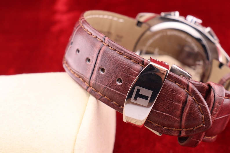 Puede incluir: Primer plano de una correa de reloj de cuero marr&oacute;n con una hebilla de acero inoxidable. La correa del reloj tiene un patr&oacute;n texturizado, similar al de un cocodrilo. La esfera del reloj es visible en el fondo. La correa del reloj est&aacute; sobre una superficie blanca sobre un fondo rojo.
