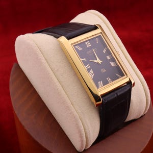 Puede incluir: Un reloj de pulsera rectangular dorado con esfera negra y números romanos. El reloj tiene una correa de cuero negro y se exhibe sobre un cojín color crema. La esfera del reloj dice "SEIKO" y "SB".