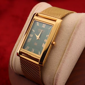 K&ouml;nnte beinhalten: Rechteckige goldfarbene Armbanduhr mit gr&uuml;nem Zifferblatt und goldenem Mesh-Armband. Das Zifferblatt der Uhr hat r&ouml;mische Ziffern und goldene Zeiger. Die Uhr wird auf einem cremefarbenen Kissen vor rotem Hintergrund pr&auml;sentiert.