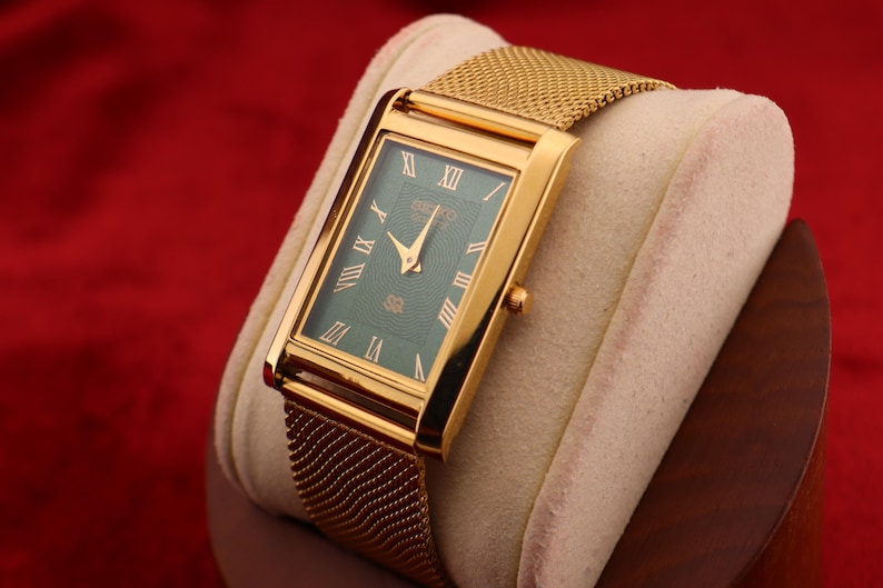 K&ouml;nnte beinhalten: Rechteckige goldfarbene Armbanduhr mit gr&uuml;nem Zifferblatt und goldenem Mesh-Armband. Das Zifferblatt der Uhr hat r&ouml;mische Ziffern und goldene Zeiger. Die Uhr wird auf einem cremefarbenen Kissen vor rotem Hintergrund pr&auml;sentiert.