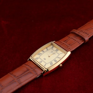 May include: A gold-toned rectangular wristwatch with a cream-colored face and Roman numeral hour markers. The watch has a brown leather band with a crocodile pattern. The watch is set against a dark red background.