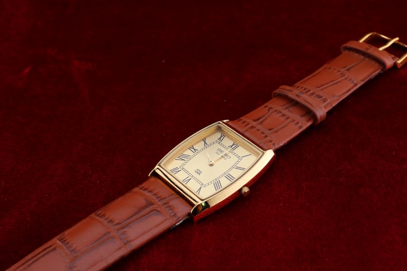 May include: A gold-toned rectangular wristwatch with a cream-colored face and Roman numeral hour markers. The watch has a brown leather band with a crocodile pattern. The watch is set against a dark red background.