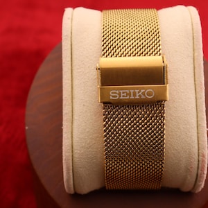 K&ouml;nnte beinhalten: Ein goldfarbenes Mesh-Uhrenarmband mit einer Schlie&szlig;e mit dem Markennamen "SEIKO". Das Armband wird auf einem cremefarbenen Kissen vor rotem Hintergrund pr&auml;sentiert. Das Uhrenarmband besteht aus feinem, gewebtem Metall.
