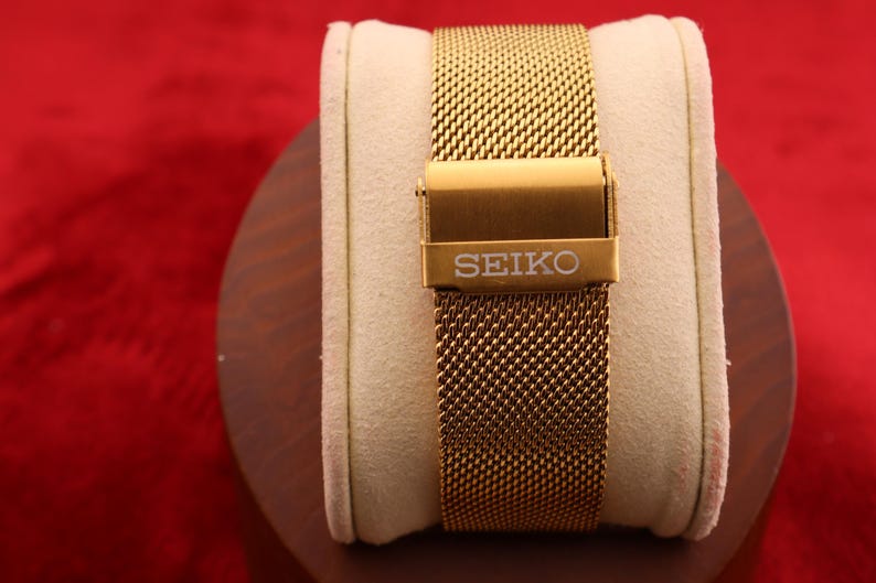 K&ouml;nnte beinhalten: Ein goldfarbenes Mesh-Uhrenarmband mit einer Schlie&szlig;e mit dem Markennamen "SEIKO". Das Armband wird auf einem cremefarbenen Kissen vor rotem Hintergrund pr&auml;sentiert. Das Uhrenarmband besteht aus feinem, gewebtem Metall.