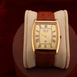 May include: A gold-toned Seiko Quartz watch with a rectangular face and brown leather band. The watch face features Roman numerals and the Seiko logo. The watch is displayed on a cream-colored cushion.