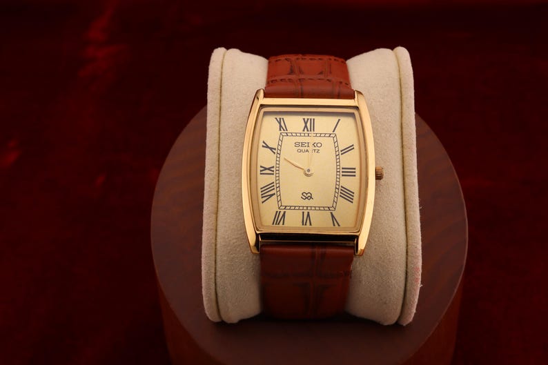 May include: A gold-toned Seiko Quartz watch with a rectangular face and brown leather band. The watch face features Roman numerals and the Seiko logo. The watch is displayed on a cream-colored cushion.