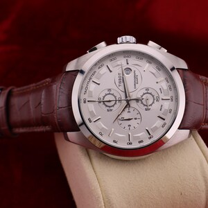 Puede incluir: Un reloj cron&oacute;grafo Tissot plateado con esfera blanca y correa de cuero marr&oacute;n. El reloj presenta m&uacute;ltiples subesferas y una escala taquim&eacute;trica. La correa tiene un patr&oacute;n de cocodrilo. El reloj se exhibe sobre una superficie color crema.