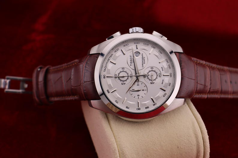 Puede incluir: Un reloj cron&oacute;grafo Tissot plateado con esfera blanca y correa de cuero marr&oacute;n. El reloj presenta m&uacute;ltiples subesferas y una escala taquim&eacute;trica. La correa tiene un patr&oacute;n de cocodrilo. El reloj se exhibe sobre una superficie color crema.