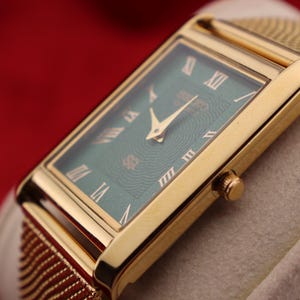 K&ouml;nnte beinhalten: Eine rechteckige Armbanduhr in Gold mit gr&uuml;nem Zifferblatt und goldenen r&ouml;mischen Ziffern. Die Uhr hat ein goldenes Mesh-Armband und eine goldene Krone. Die Uhr wird auf einer hellen Oberfl&auml;che vor rotem Hintergrund pr&auml;sentiert.