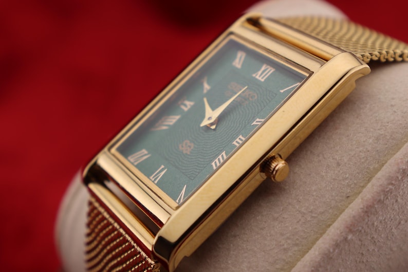 K&ouml;nnte beinhalten: Eine rechteckige Armbanduhr in Gold mit gr&uuml;nem Zifferblatt und goldenen r&ouml;mischen Ziffern. Die Uhr hat ein goldenes Mesh-Armband und eine goldene Krone. Die Uhr wird auf einer hellen Oberfl&auml;che vor rotem Hintergrund pr&auml;sentiert.