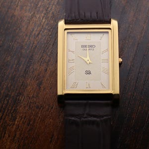 May include: A gold-toned rectangular Seiko Quartz watch with a cream-colored face and black leather band. The watch face features Roman numerals and the Seiko logo. The watch is displayed on a dark brown wooden surface.