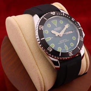 May include: A Seiko automatic sports diver's watch with a black bezel and a black rubber strap. The watch face is teal with white hands and markers. The watch is displayed on a cream-colored cushion.