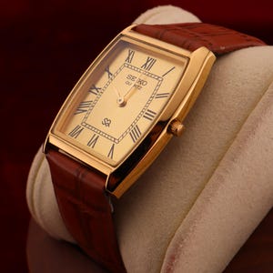 May include: A gold-toned Seiko Quartz watch with a rectangular face and brown leather strap. The watch face features Roman numerals and the Seiko logo. The watch is displayed on a cream-colored cushion against a dark red background.