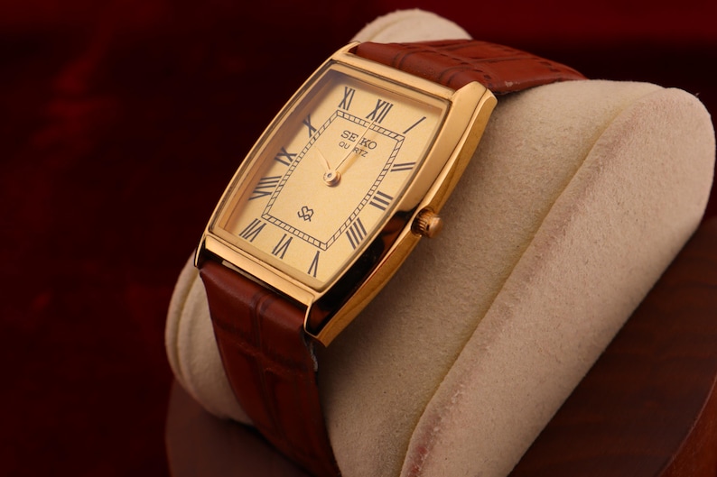 May include: A gold-toned Seiko Quartz watch with a rectangular face and brown leather strap. The watch face features Roman numerals and the Seiko logo. The watch is displayed on a cream-colored cushion against a dark red background.