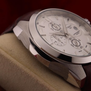 Puede incluir: Primer plano de un reloj de pulsera plateado con esfera blanca y tres subesferas. El reloj tiene una correa de cuero marr&oacute;n. El reloj est&aacute; sobre una superficie beige sobre un fondo rojo oscuro.