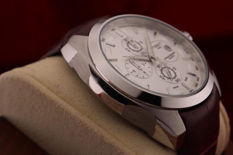 Puede incluir: Primer plano de un reloj de pulsera plateado con esfera blanca y tres subesferas. El reloj tiene una correa de cuero marr&oacute;n. El reloj est&aacute; sobre una superficie beige sobre un fondo rojo oscuro.