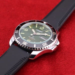 Puede incluir: Un reloj de pulsera de acero inoxidable con bisel negro y correa de caucho negra. La esfera del reloj es verde con n&uacute;meros y agujas blancas. El reloj est&aacute; inclinado sobre una superficie roja.